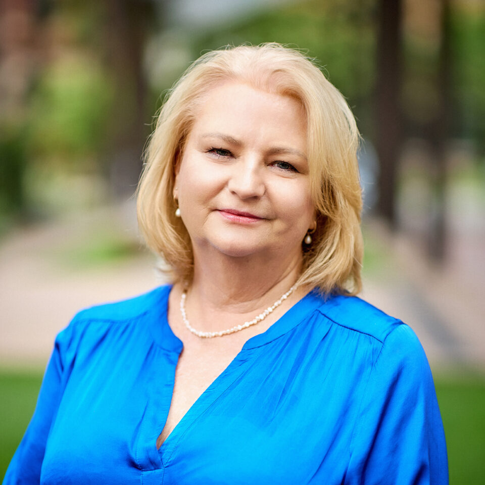 Michelle Teis Headshot - Michellle wears a blue blouse with a white necklace and earrings.