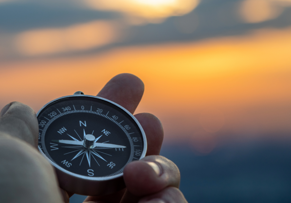 A persons hand holding a compass showing North south east and west against a nice sunset