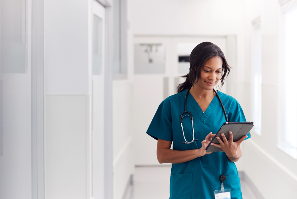 "Healthcare professional in medical scrubs with a stethoscope, standing in a hospital hallway and reviewing information on a tablet device."