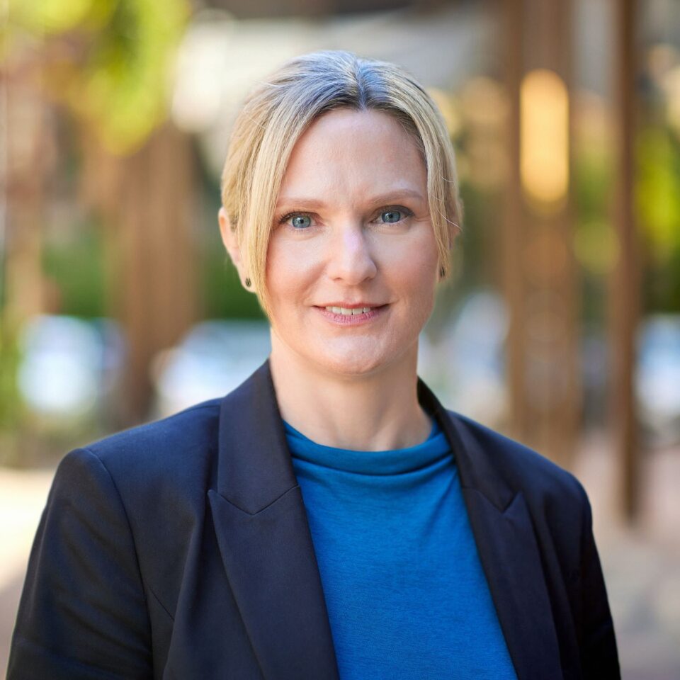 Jane Brimacombe Headshot - Jane wears a blue shirt with a dark jacket with her hair up.