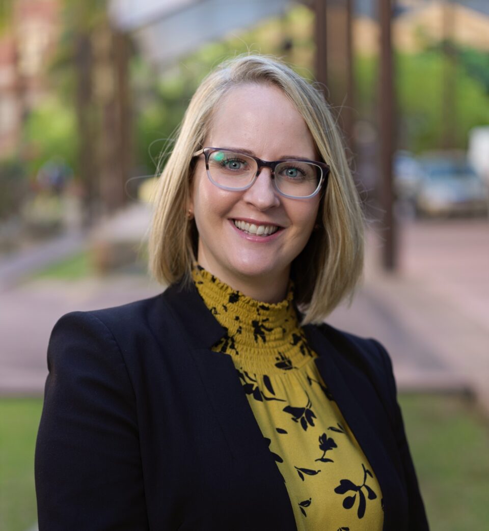 Jen-Coulthard-Headshot - Jen wears a yellow and black print top with a black jacket. She has blonde short hair and wears glasses.