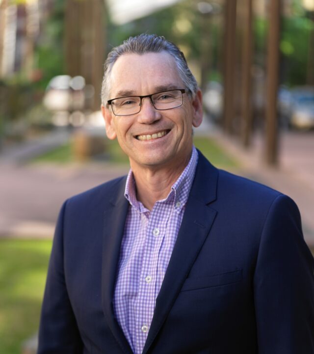 Jon O'Brien Headshot - Jon has a checkered shirt and blue jacket, he wears glasses.