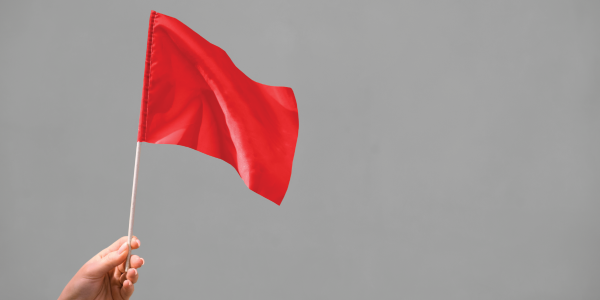 "Hand holding a small red flag on a stick against a plain grey background, symbolising a warning or alert."
