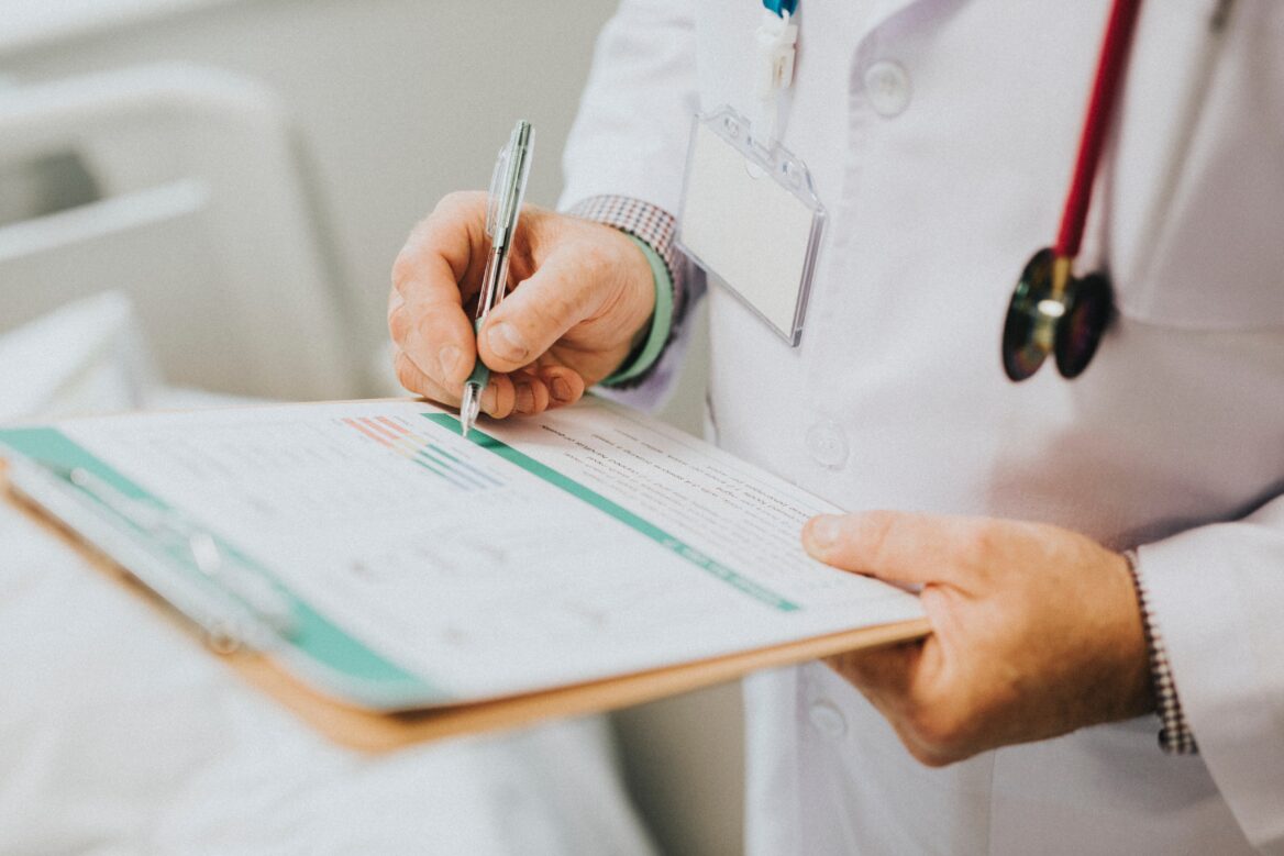 "Medical professional in a white coat with a stethoscope and ID badge, writing on a clipboard with forms or charts, representing healthcare documentation and patient care."