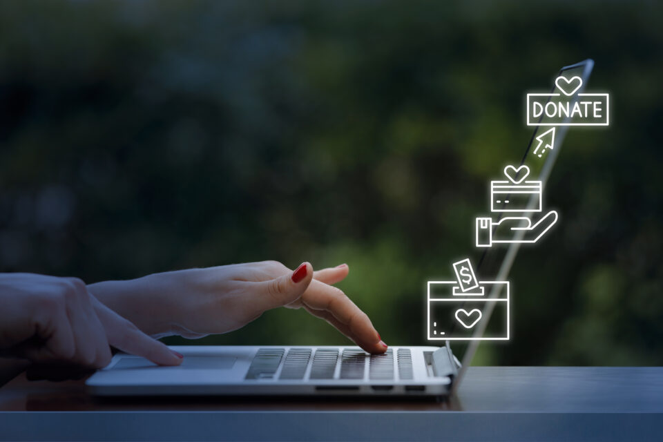 "Hand interacting with a laptop, with digital icons above showing a wallet, donation box, and the word 'DONATE', symbolising online charitable contributions."