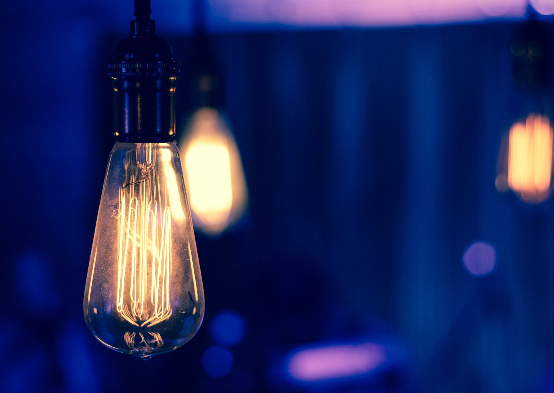 "Close-up of an illuminated vintage Edison light bulb with visible filaments, set against a blurred background of additional bulbs and a moody blue-purple glow."
